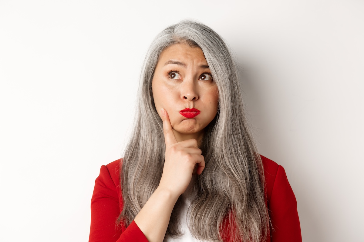 Close up of indecisive asian senior woman making choice, pouting and looking thoughtful at upper left corner, standing against white background
